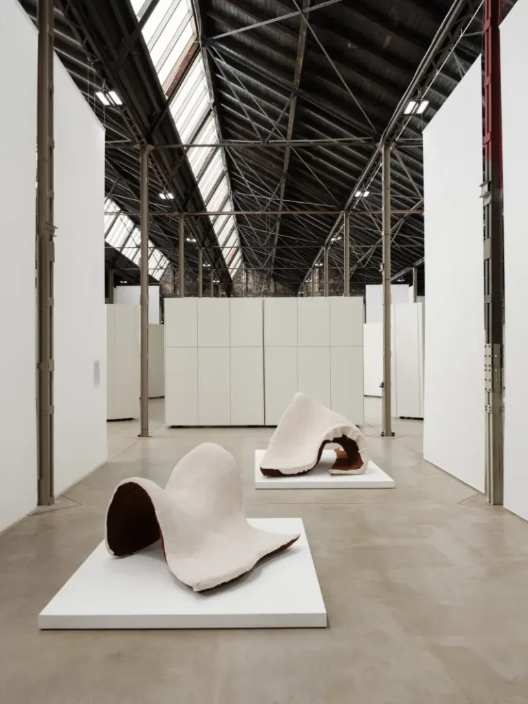 Two white, irregular sculptural forms with dark interiors sit on plinths in a large industrial gallery with a steel roof