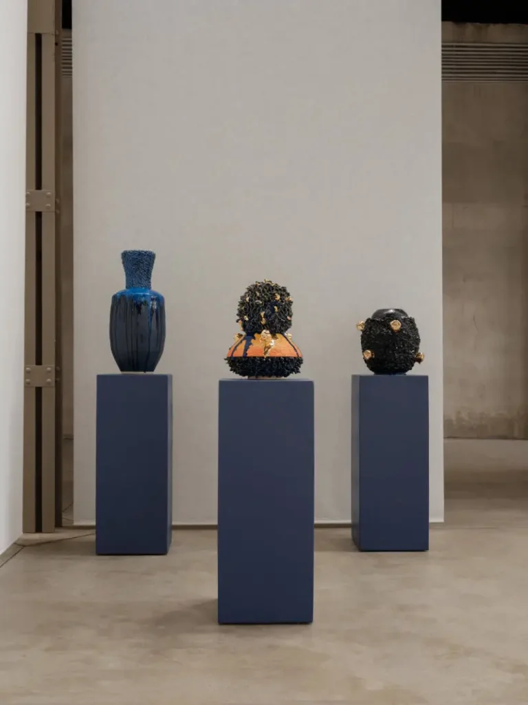 Three sculptural objects displayed on dark blue pedestals in a minimalist gallery: a blue vase and two black textured forms
