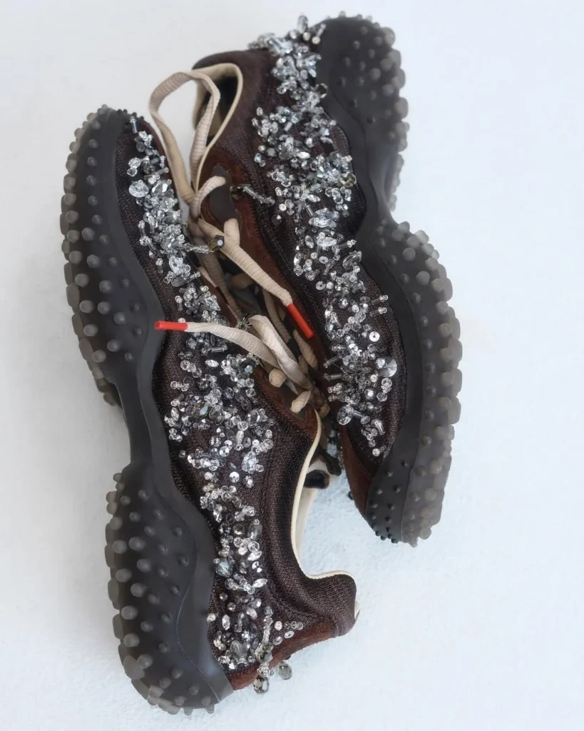 Pair of brown low-profile sneakers covered in silver embellishments, with black soles lined by rounded rubber studs