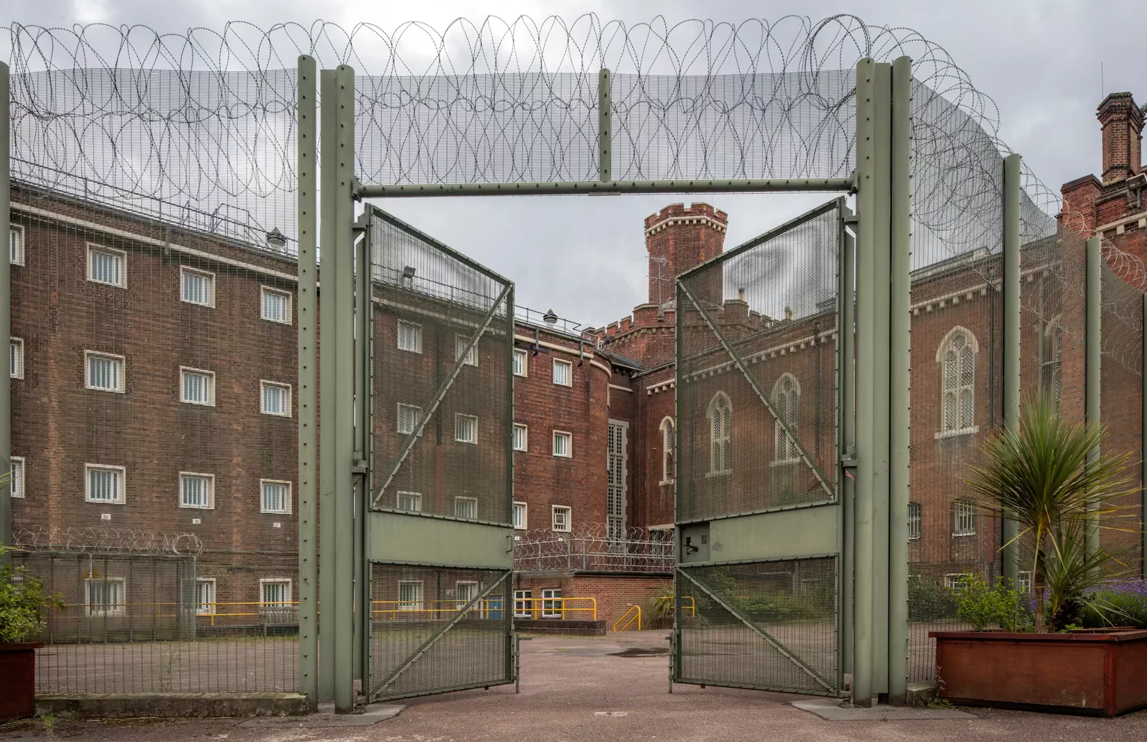 Open security gates topped with razor wire reveal red-brick prison buildings and barred windows under a gray sky