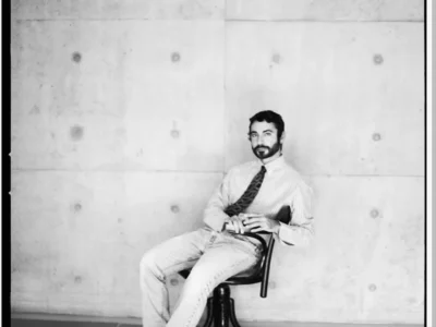 Dario Vitale seated in a black chair, wearing a striped shirt, patterned tie, light jeans and black shoes against a concrete