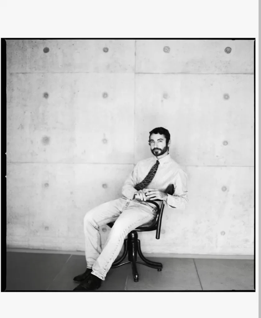 Dario Vitale seated in a black chair, wearing a striped shirt, patterned tie, light jeans and black shoes against a concrete