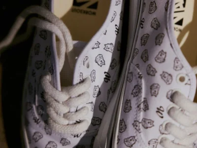 Close-up of white Vans shoe covered in small cartoon face and letter prints, with white laces and logo insoles on a dark