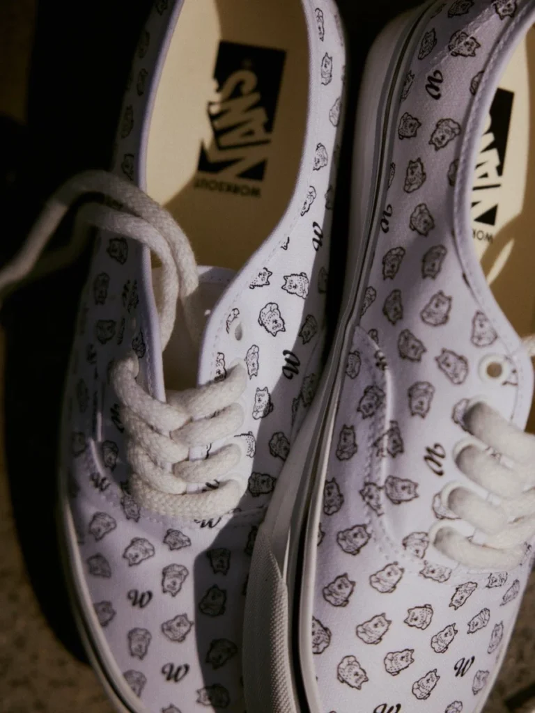 Close-up of white Vans shoe covered in small cartoon face and letter prints, with white laces and logo insoles on a dark