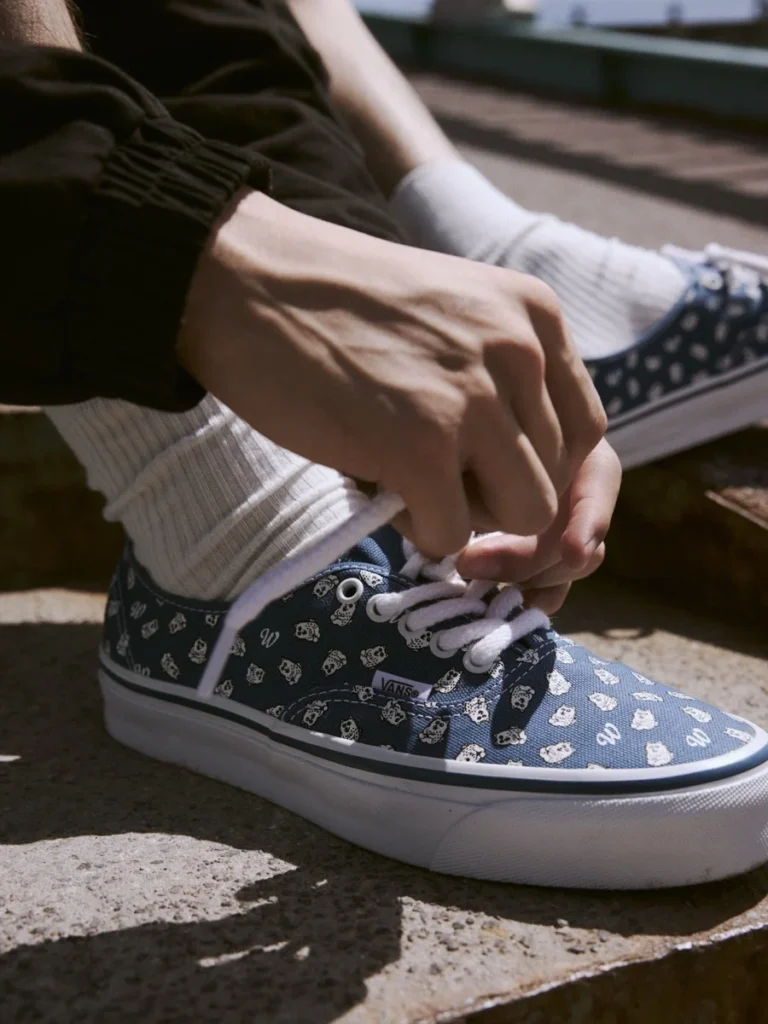 Person in white ribbed socks ties white laces on blue Vans sneakers patterned with small skulls and logos
