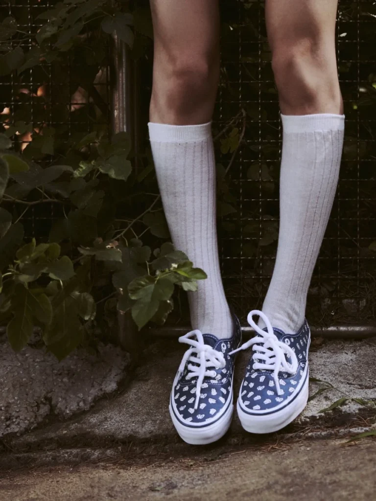 Lower legs in ribbed white knee socks and navy patterned lace-up sneakers, standing by a leafy fence