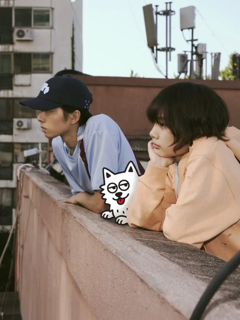 Two people lean on a rooftop ledge, with a cartoon cat sticker between them and city buildings and antennas behind