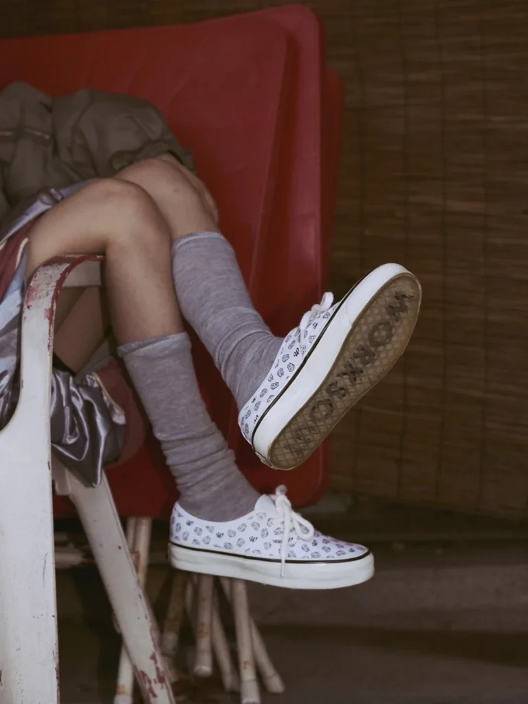 Person’s crossed legs in gray knee-high socks and white patterned sneakers, seated on a red chair beside a wooden wall