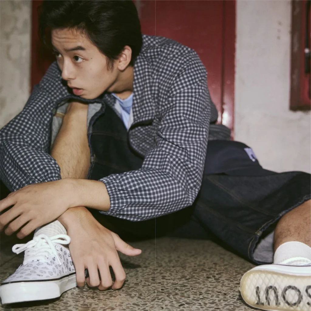 Person seated on a terrazzo floor wearing a blue checked jacket, black shorts, white patterned sneakers, and socks