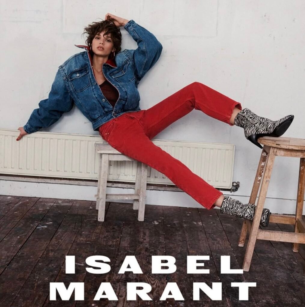 Model in a denim jacket and red pants reclines across stools, wearing zebra-print ankle boots in a loft setting