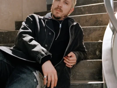 Glenn Martens sits on dark stone stairs wearing a black leather jacket and black jeans, with rings on his fingers