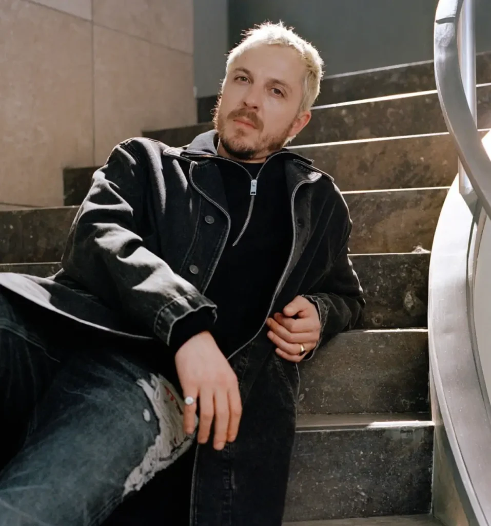 Glenn Martens sits on dark stone stairs wearing a black leather jacket and black jeans, with rings on his fingers