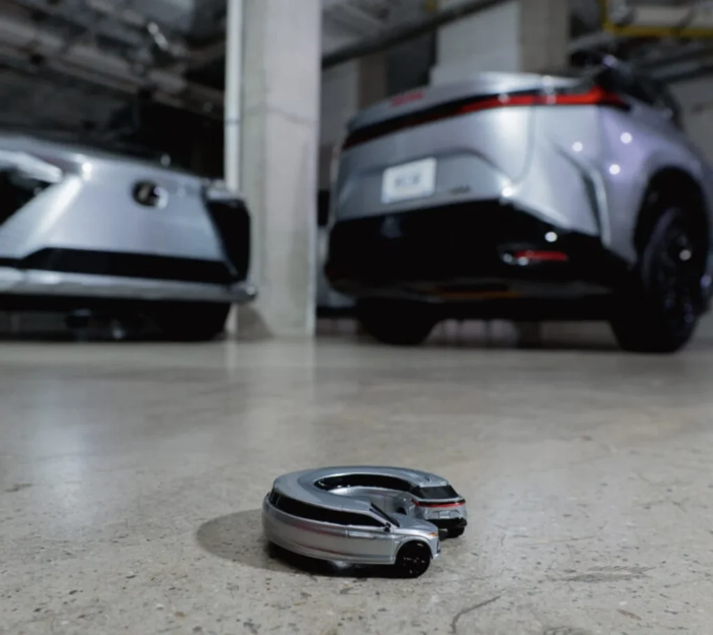 Small silver car sculpture on a concrete floor, with two full-size silver vehicles blurred in the background