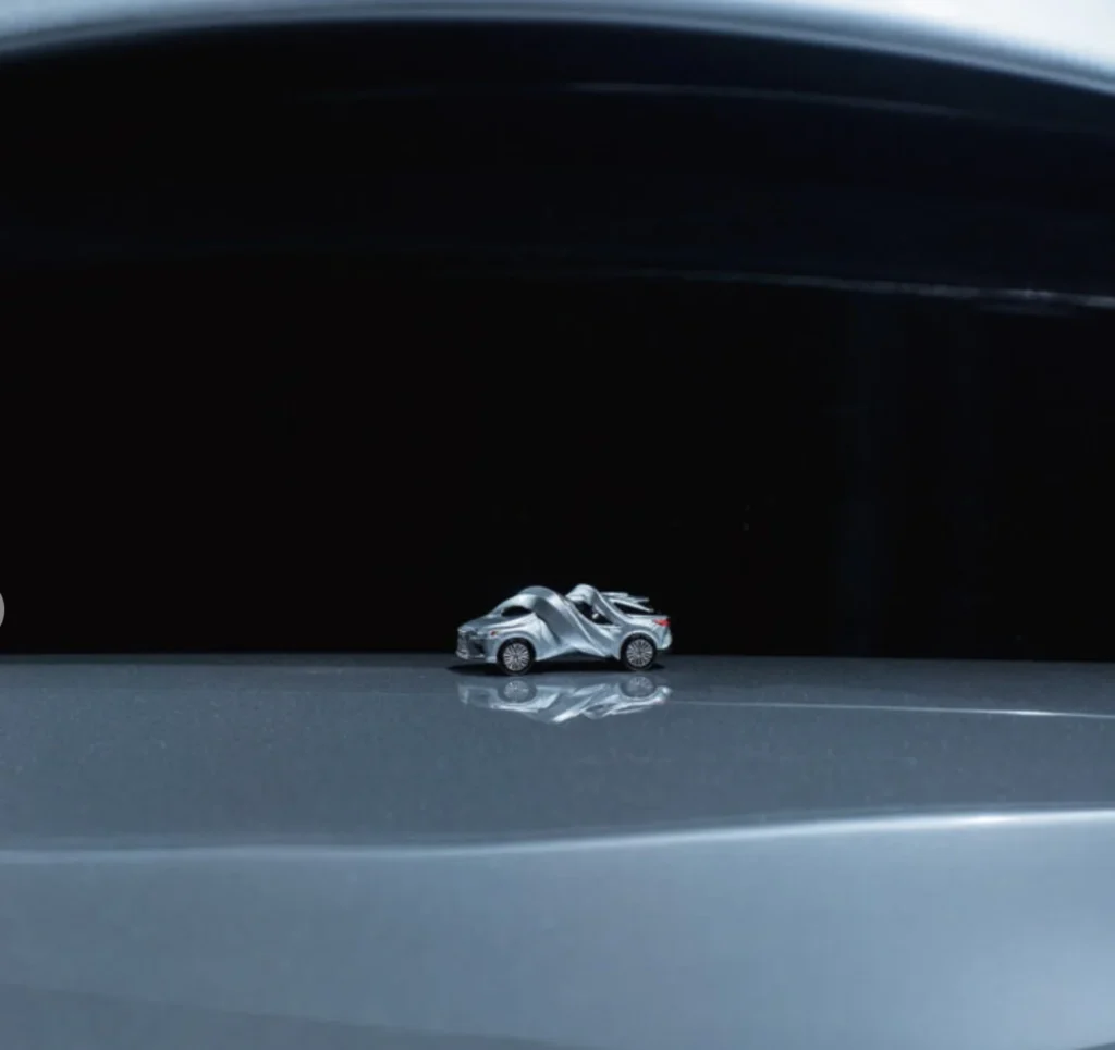 Small sculptural car made from two silver vehicles, displayed on a reflective platform against a black background