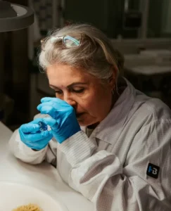 Woman in a cleanroom suit and blue gloves examines a small camera component under a magnifying lamp