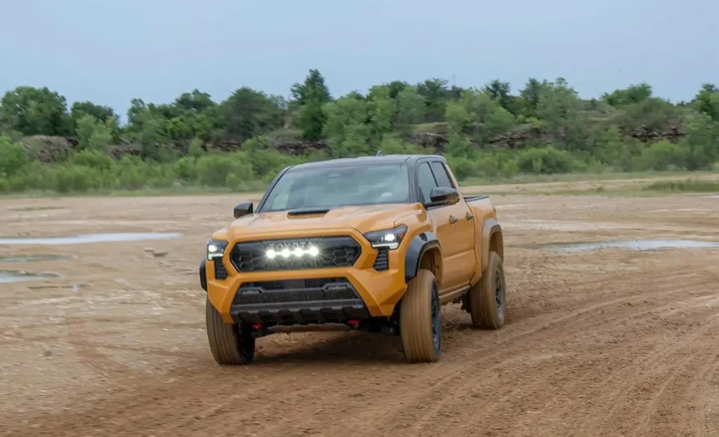 Orange Toyota pickup truck driving across a muddy off-road area, with trees and rocky terrain in the background