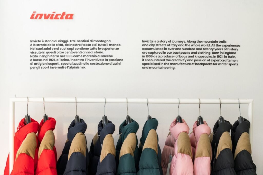 Invicta logo and bilingual brand history text above a rack of red, navy, green, pink, and black padded jackets