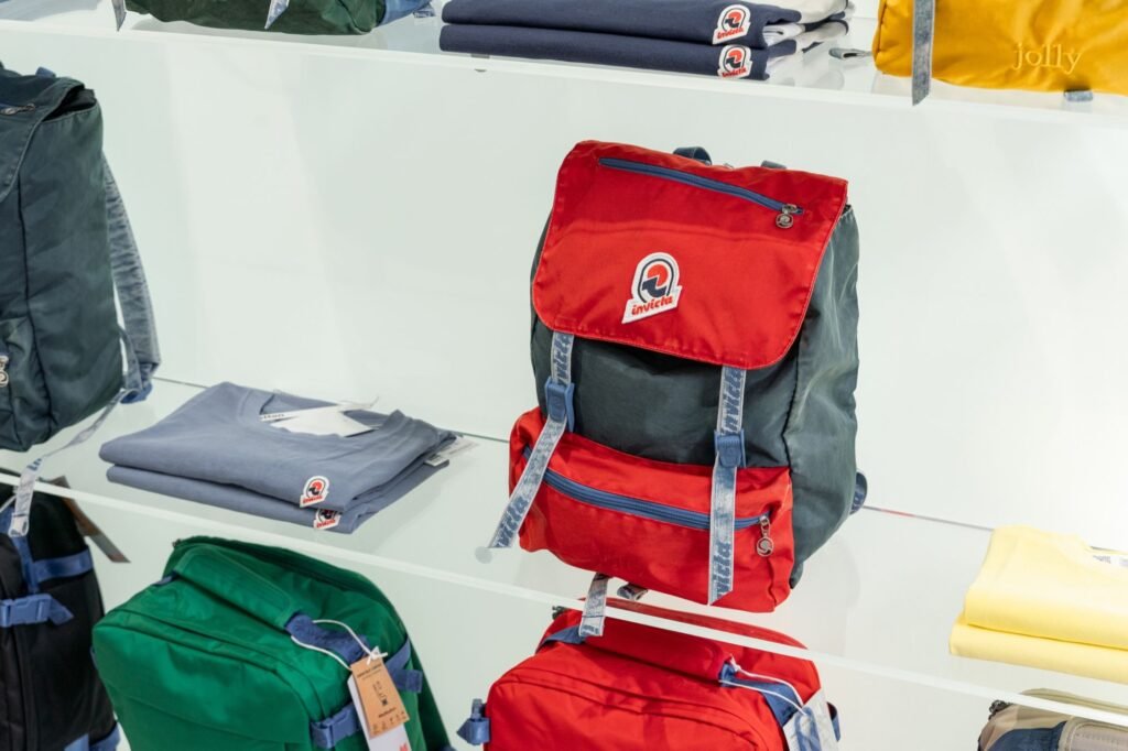 Red and dark gray Invicta backpack with blue straps displayed among folded clothing and other bags on white shelves