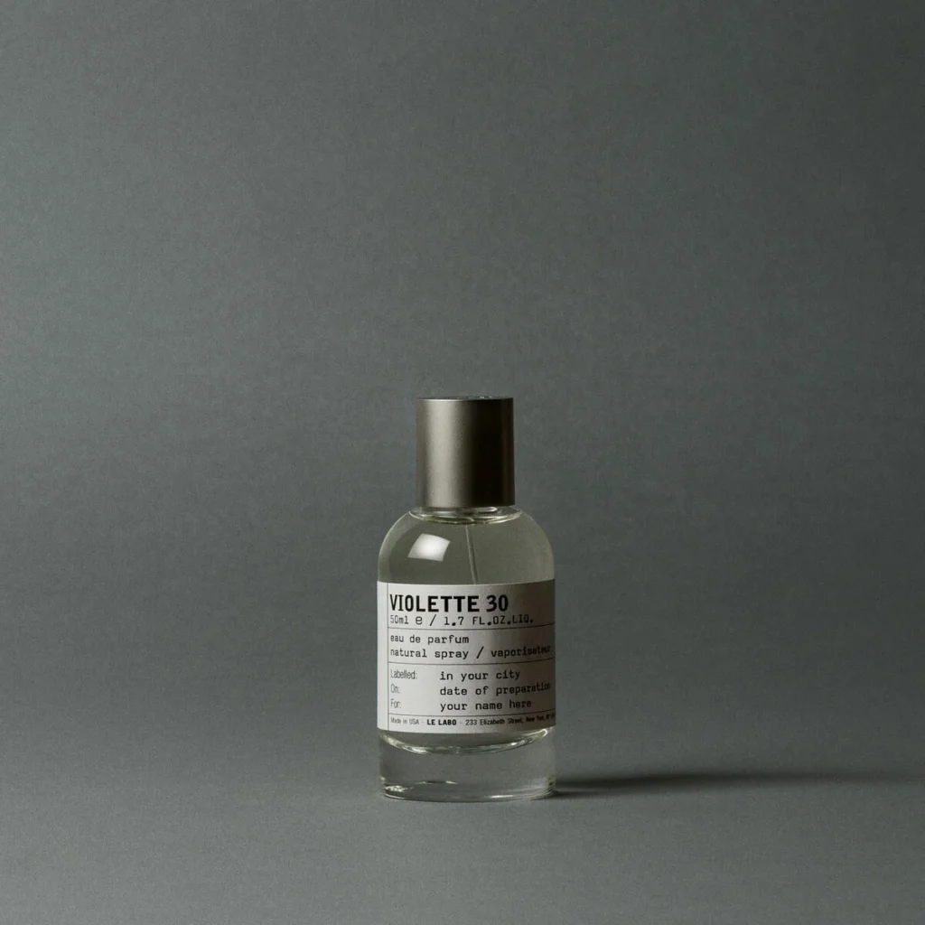 Clear glass Violette 30 perfume bottle with metallic cap and white label on a gray surface against a gray backdrop