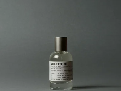 Clear glass Violette 30 perfume bottle with metallic cap and white label on a gray surface against a gray backdrop