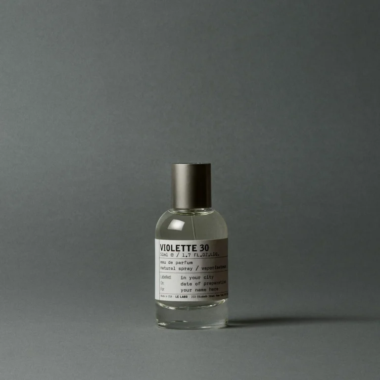 Clear glass Violette 30 perfume bottle with metallic cap and white label on a gray surface against a gray backdrop