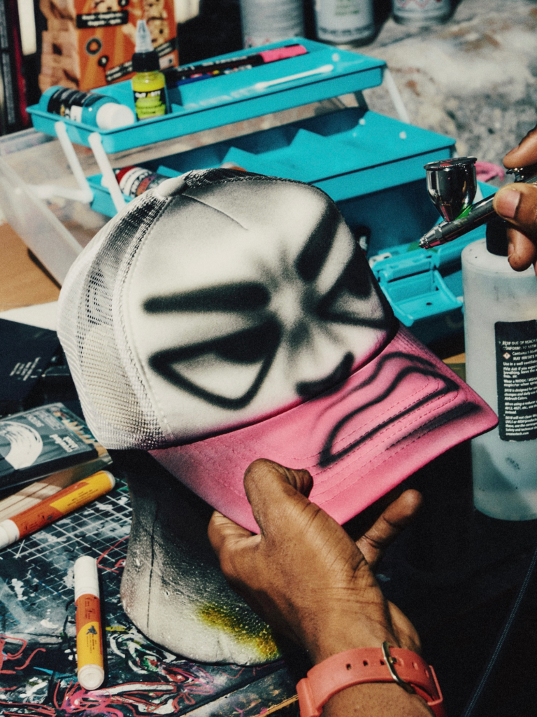 A hand holds a pink-and-white mesh cap with black airbrushed lettering beside an airbrush and art supplies