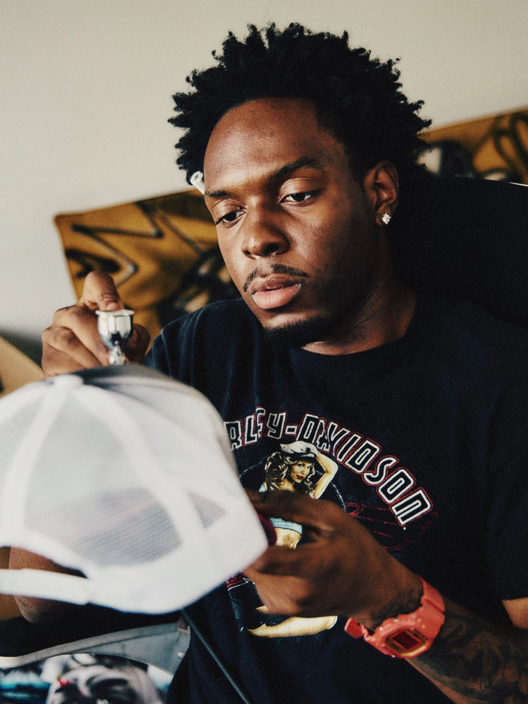 Man with short afro holds a tattoo machine over a client’s white cap, wearing a dark Playboy shirt and red watch