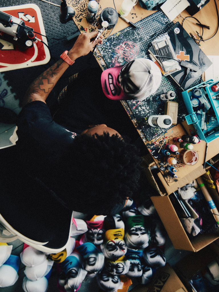Person in a pink-and-white cap paints at a cluttered craft table surrounded by markers, bottles, tools and masks