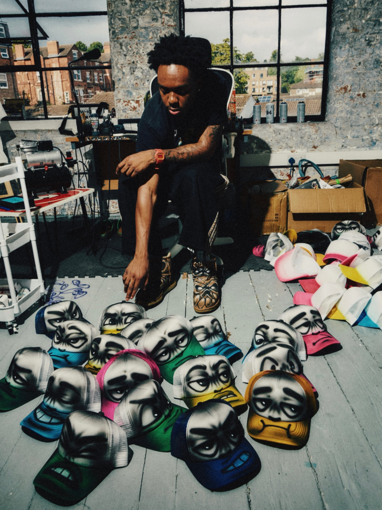 Tattooed person in black clothing crouches among colorful caps spray-painted with cartoon faces in a sunlit studio