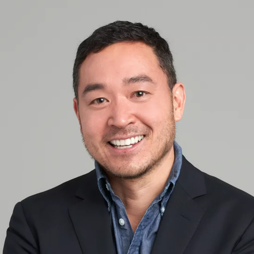 Smiling man with short dark hair wearing a black blazer and blue button-down shirt against a gray background