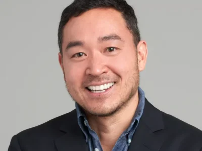Smiling man with short dark hair wearing a black blazer and blue button-down shirt against a gray background