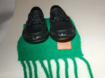 Pair of black leather loafers with braided detailing on a green fringed scarf, against a white background