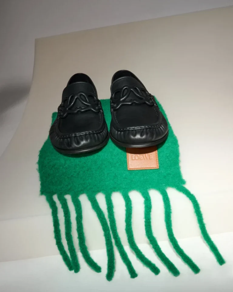 Pair of black leather loafers with braided detailing on a green fringed scarf, against a white background