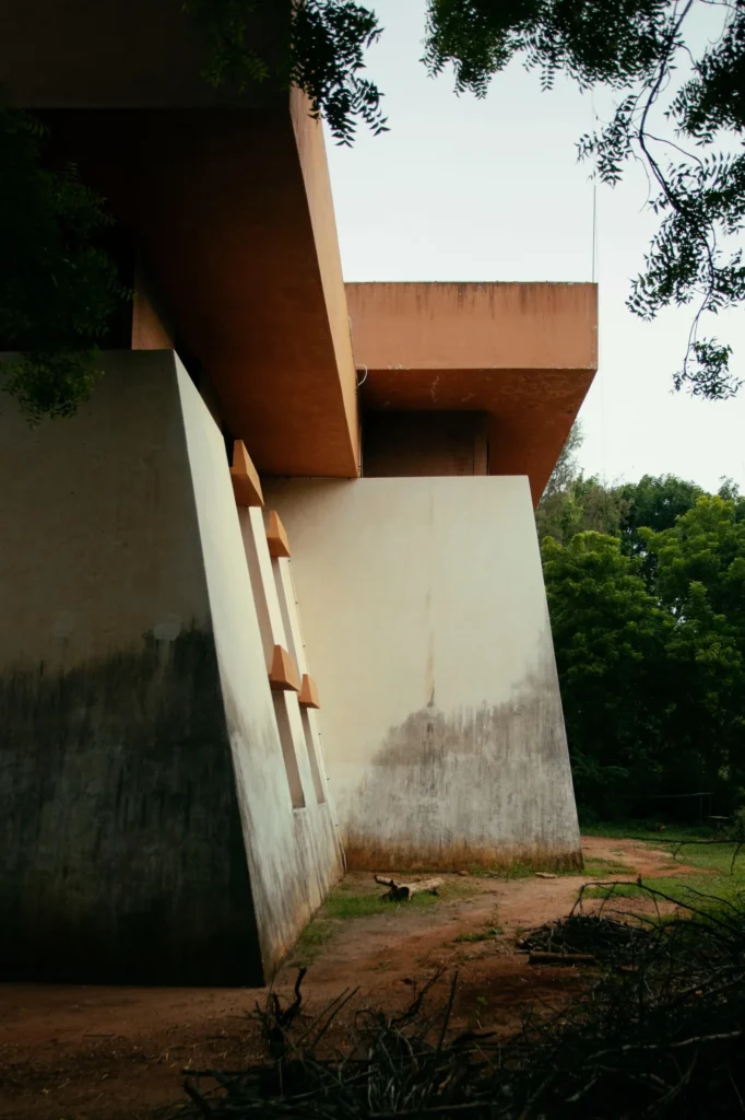 Angular concrete building with rust-colored cantilevered roofs, white walls, weathering, trees, and dirt ground
