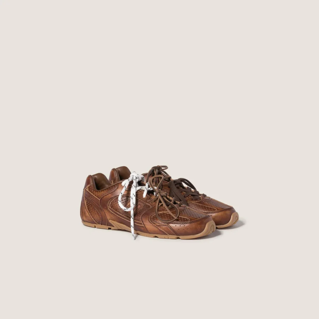 Pair of low-profile brown leather sneakers with perforated panels and contrasting white and brown laces on a beige background