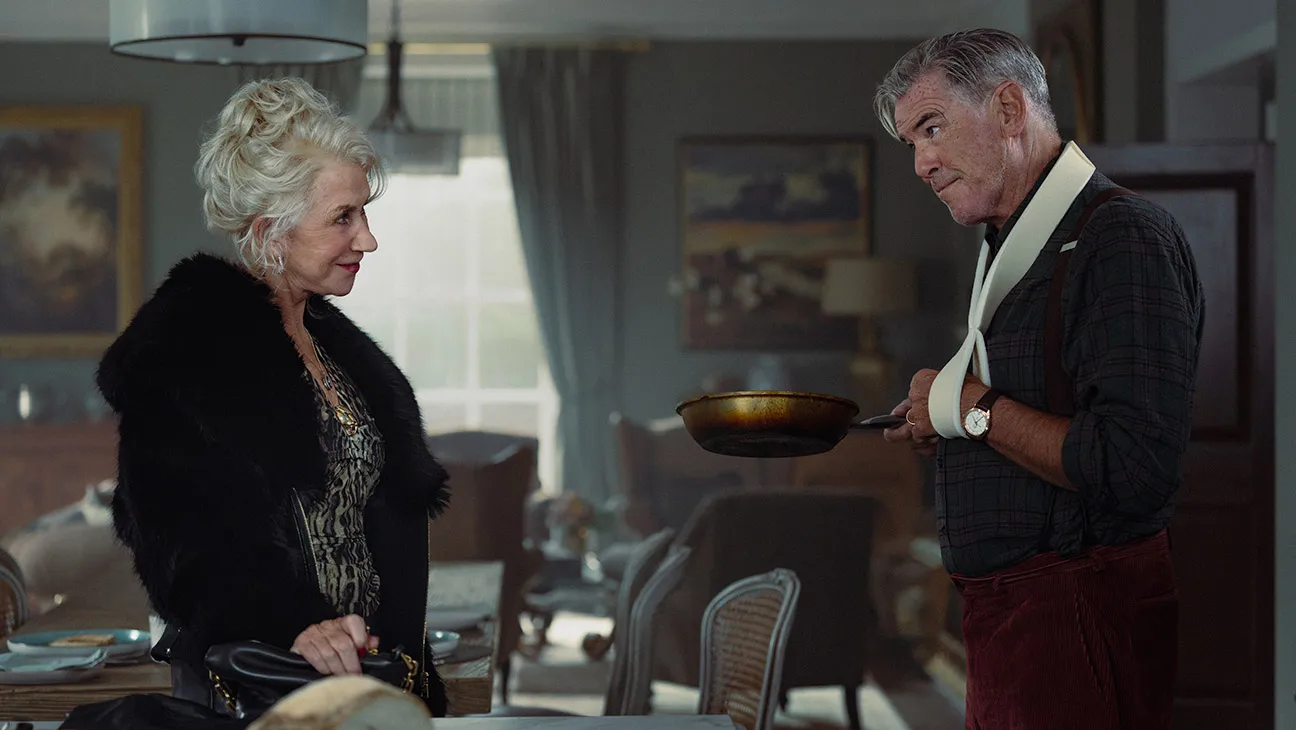 Right side Pierce Brosnan left arm in a sling raised holding a frypan and Left side Helen Mirren in short-tailed Black Fur Jacket, right hand press on purse kitchen table, living room sil 
