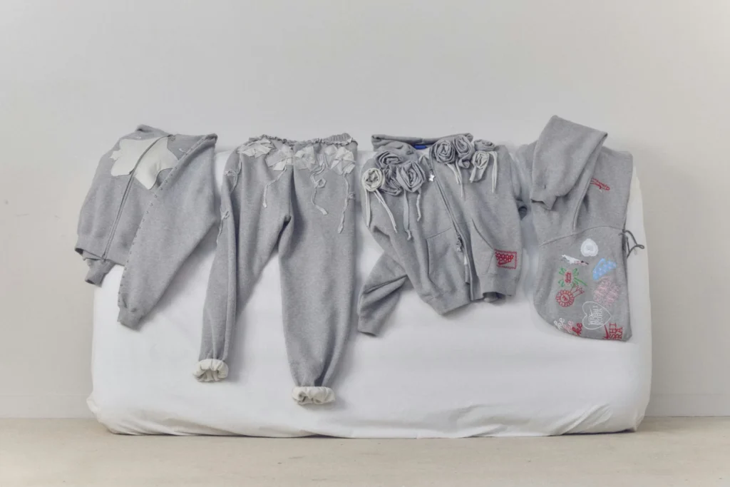 Gray embellished clothing arranged on a white mattress, including a cardigan, pants, floral hoodie, and embroidered bag
