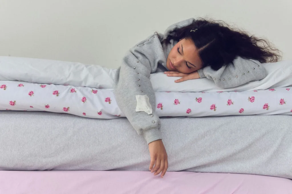 Person lying across a bed with eyes closed, wearing a gray studded sweatshirt; one arm hangs over pink bedding