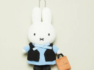 White rabbit plush keychain wearing a blue dress and black vest, holding an orange United Arrows bag against a pale