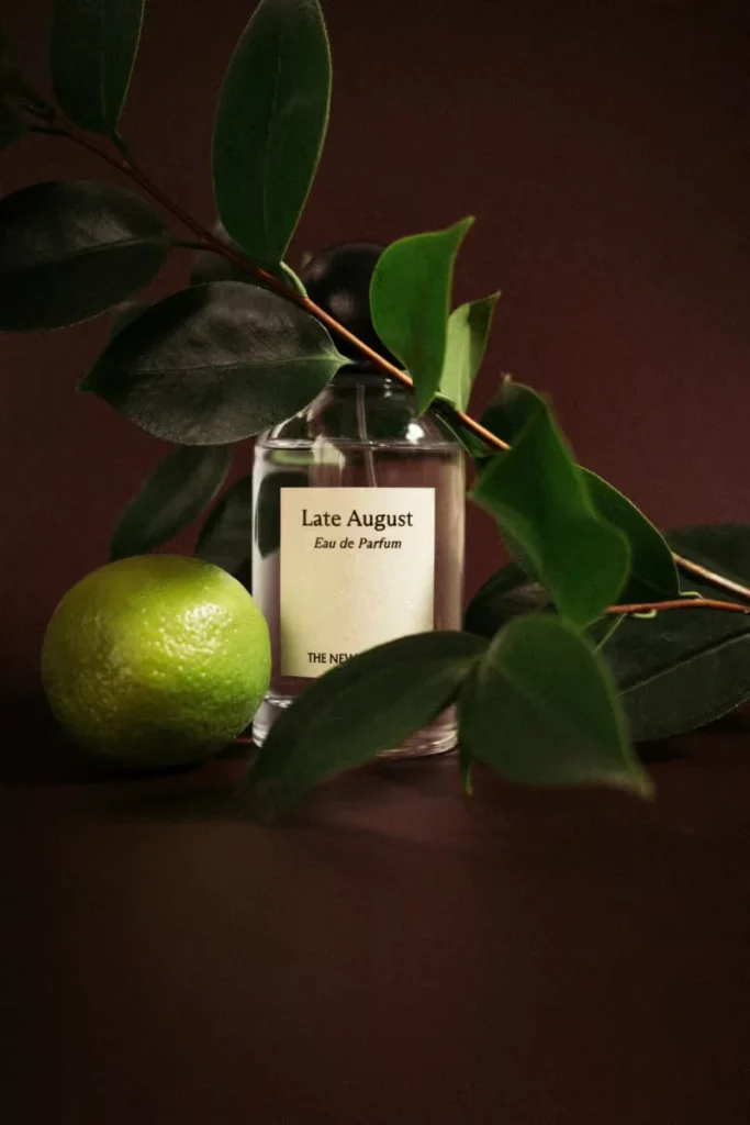 Glass perfume bottle labeled “Late August Eau de Parfum” among green leaves and a lime on a dark brown surface