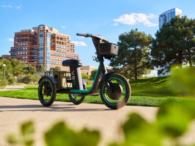 Black-and-teal electric trike with a front basket and rear cargo area parked on a park path, with city buildings behind