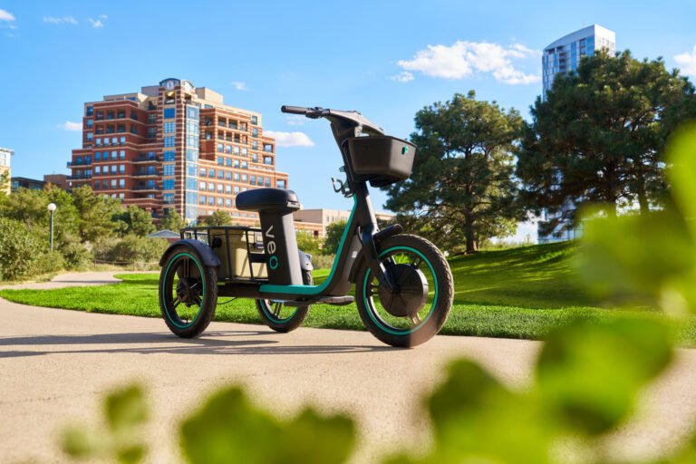 Black-and-teal electric trike with a front basket and rear cargo area parked on a park path, with city buildings behind