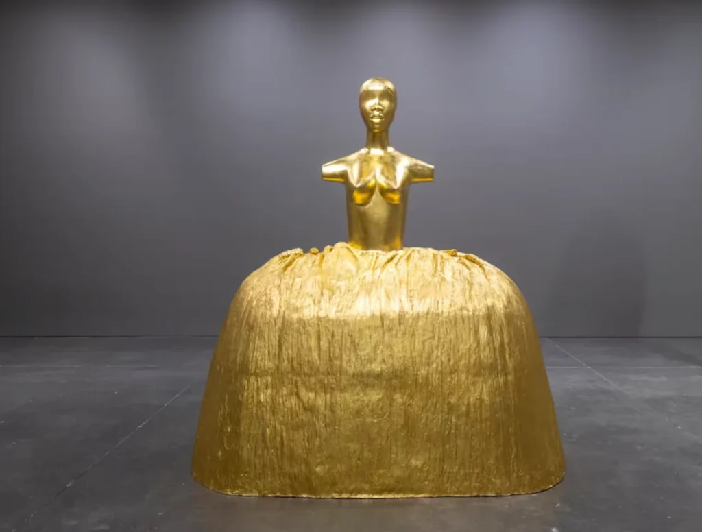 Golden sculpture figure with a human torso emerging from a large textured bell-shaped gold base in a gallery