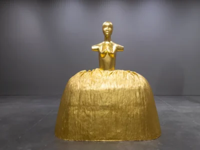 Golden sculpture figure with a human torso emerging from a large textured bell-shaped gold base in a gallery