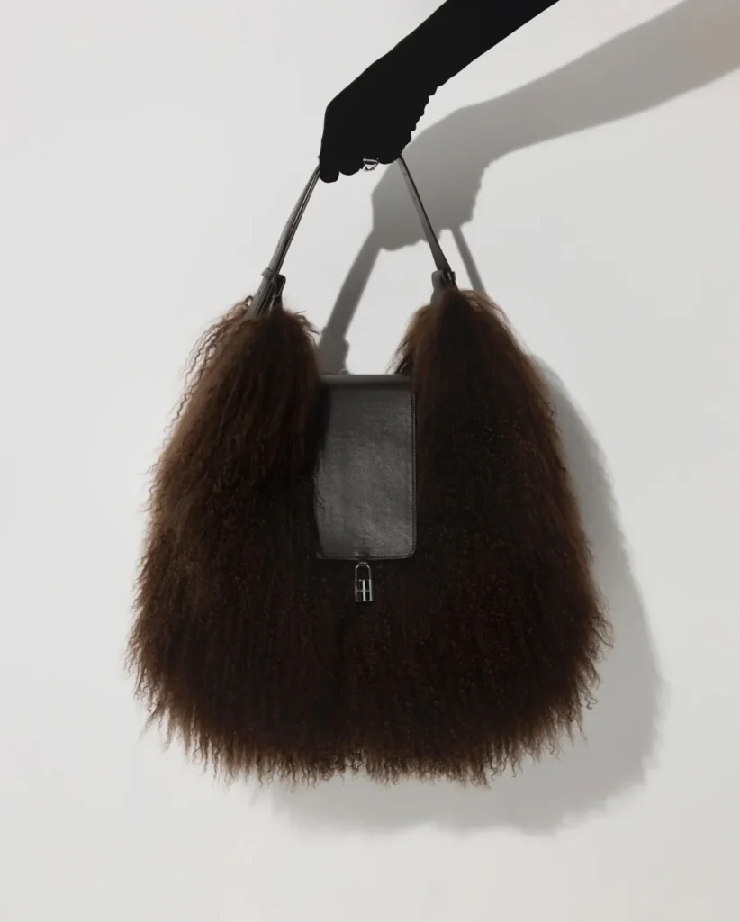 Black leather handbag with a rectangular flap and large dark brown furry sides, held against a white wall casting a shadow