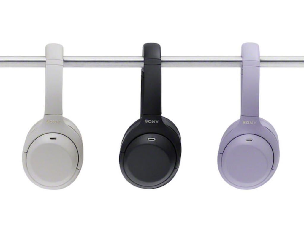 Three Sony over-ear headphones hang from a metal bar: white, black, and pale lavender models against a white background