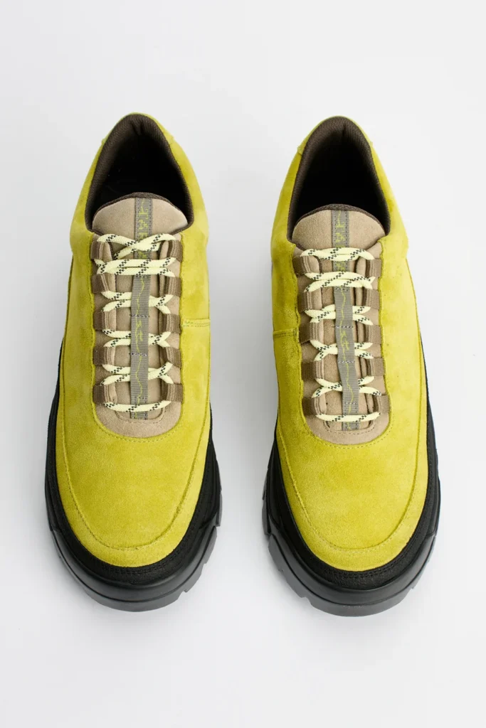 Top-down view of lime-green suede sneakers with cream rope laces, tan panels, black trim and chunky soles on a white