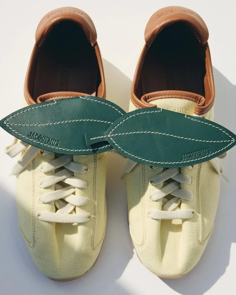 Pair of cream low-top sneakers with white laces, brown padded collars, and oversized green leather Jacquemus tongues on a