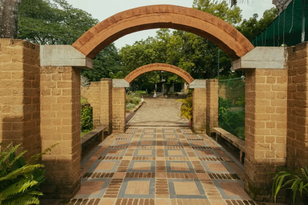 Sand-colored brick gateway with two arched openings frames a tiled walkway through lush greenery; a person stands in the