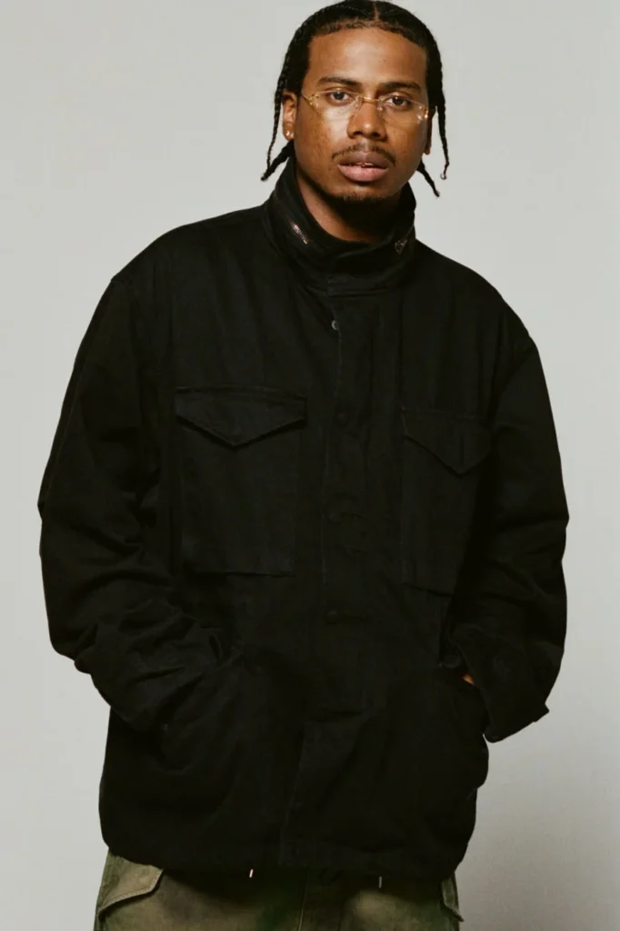 Person with braided hair and glasses wearing a black field jacket with flap pockets, hands in pockets, against a gray studio